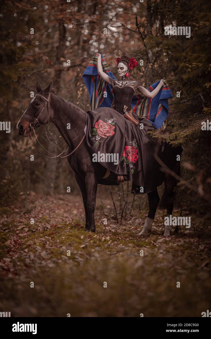 Giovane donna vestita come il simbolo messicano del giorno della calavera morta in un vestito nero che si posa in la foresta con un cavallo Foto Stock