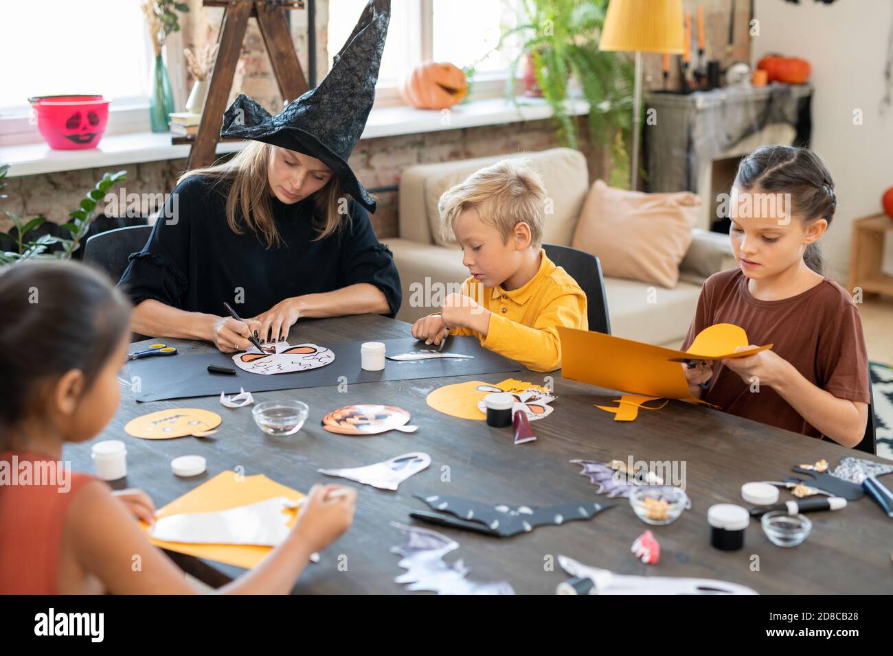 Concentrato giovane insegnante d'arte in streghe cappello seduto a tavola E disegno Halloween immagini con i capretti mentre preparando le decorazioni di carta Foto Stock