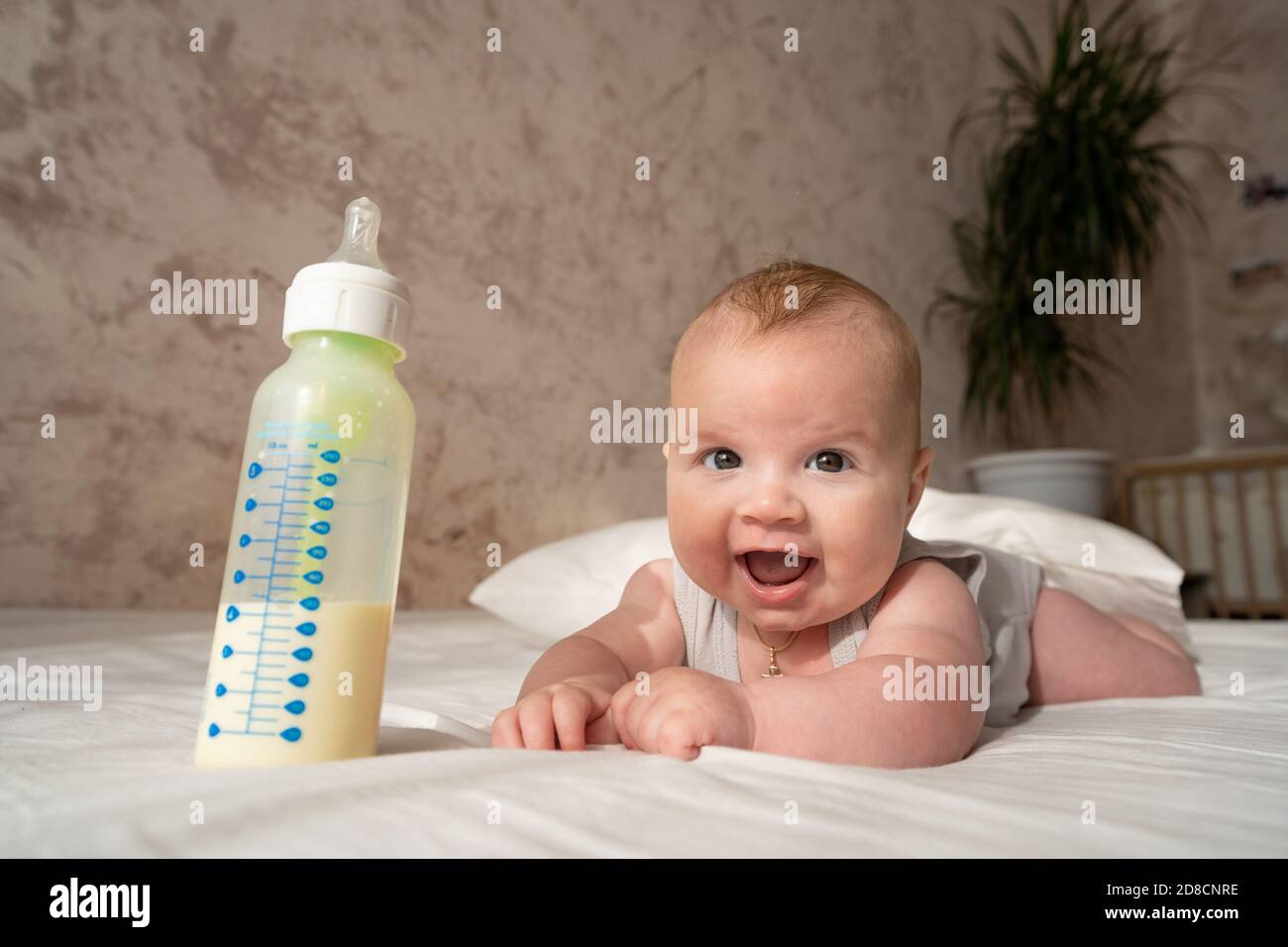 felice bambino di tre mesi giace su un letto bianco accanto a una bottiglia di latte Foto Stock
