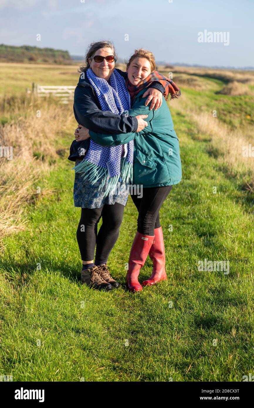 Madre e figlia adulta che camminano insieme vicino al fiume ore, Suffolk, Inghilterra, UK anteriore ritratto a lunghezza intera abbracciando Foto Stock