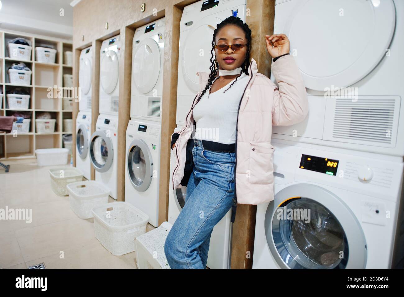 Allegra donna afroamericana in occhiali da sole vicino lavatrice nella lavanderia self-service. Foto Stock