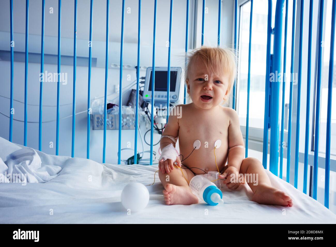 Un piccolo bambino di un anno si siede nel letto dell'ospedale e. piangi con il catetere in mano Foto Stock
