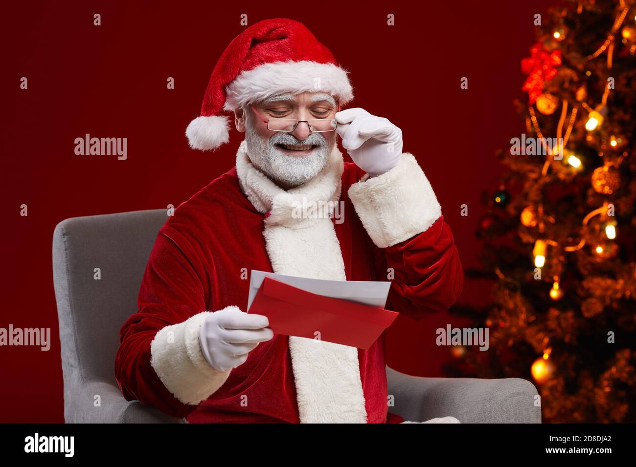 Senior Babbo Natale in costume rosso che riceve lettere dai bambini Si siede sulla poltrona vicino all'albero di Natale Foto Stock