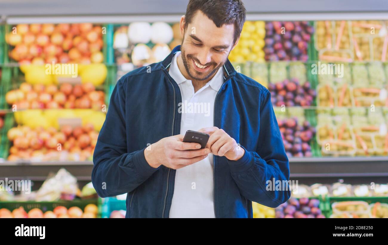 Al supermercato: Handsome Man utilizza smartphone mentre si trova nella sezione prodotti freschi del negozio. Uomo immerso in Internet Surfing sul suo Foto Stock