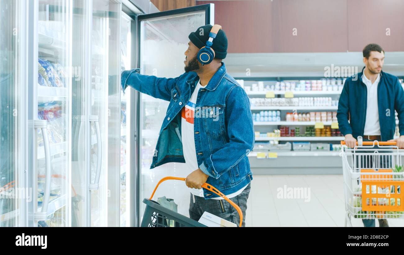 Al Supermarket: L'elegante afroamericano Guy con cuffie sceglie i prodotti nei prodotti congelati dal frigorifero e li mette in Shopping Foto Stock