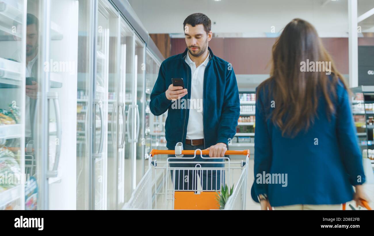 Al supermercato: Handsome Man utilizza smartphone, sorride e cammina attraverso la sezione merci surgelate. Altri clienti che acquistano in background. Foto Stock