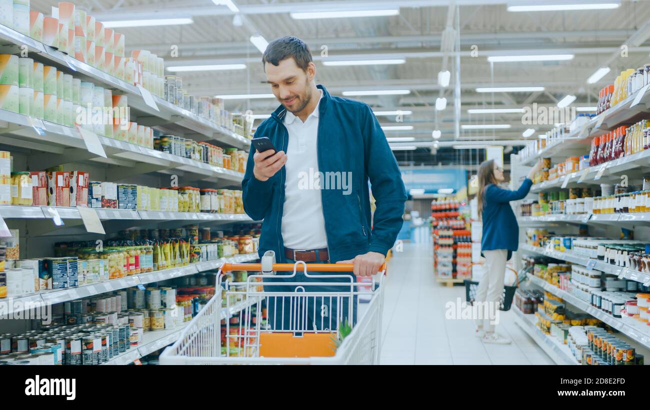 Al supermercato: Handsome Man utilizza smartphone, sorride mentre si sta in piedi alla sezione di prodotti in scatola. Ha il carrello della spesa con gli articoli sani di alimento all'interno Foto Stock