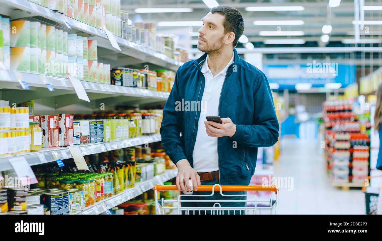 Al supermercato: L'uomo bello passa attraverso la mensola con le merci in scatola, i posti Tin possono nel suo carrello di shopping e procedere con la sua lista di shopping Foto Stock