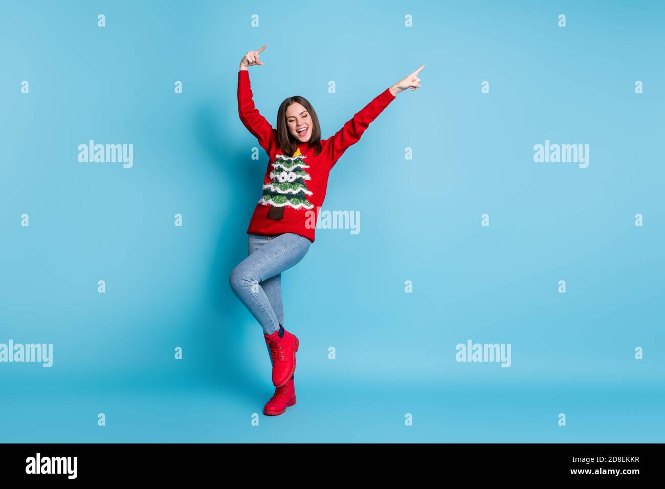 Ritratto fotografico di felice bruna ragazza che danzano sorridendo mostrando puntamento con le dita su entrambe le mani che indossano il pullover rosso di natale isolato su blu Foto Stock