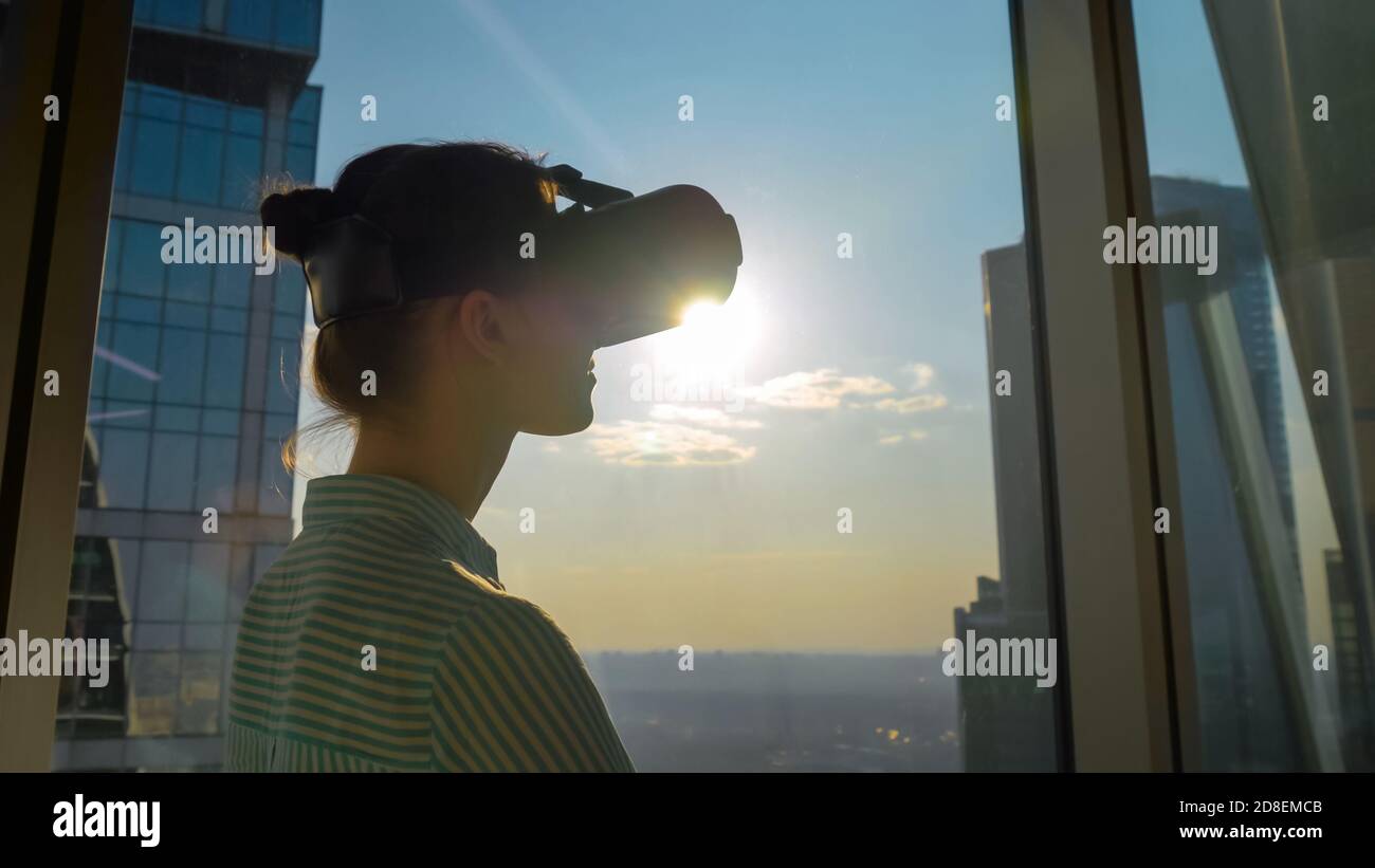 Donna che usa la cuffia per la realtà virtuale e guarda il paesaggio urbano dalla finestra panoramica dei grattacieli in ufficio. Svasatura della lente del sole. VR Foto Stock