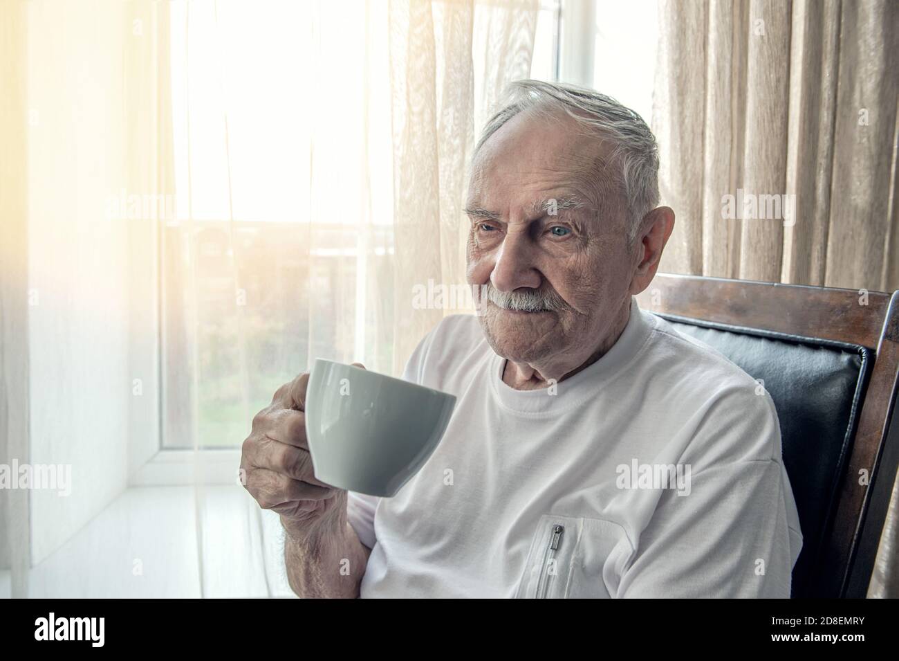 l'anziano è seduto su una sedia. Un anziano sorridente è seduto su una sedia vicino alla finestra con una tazza di caffè o tè. Ritratto, primo piano, copia spazio Foto Stock