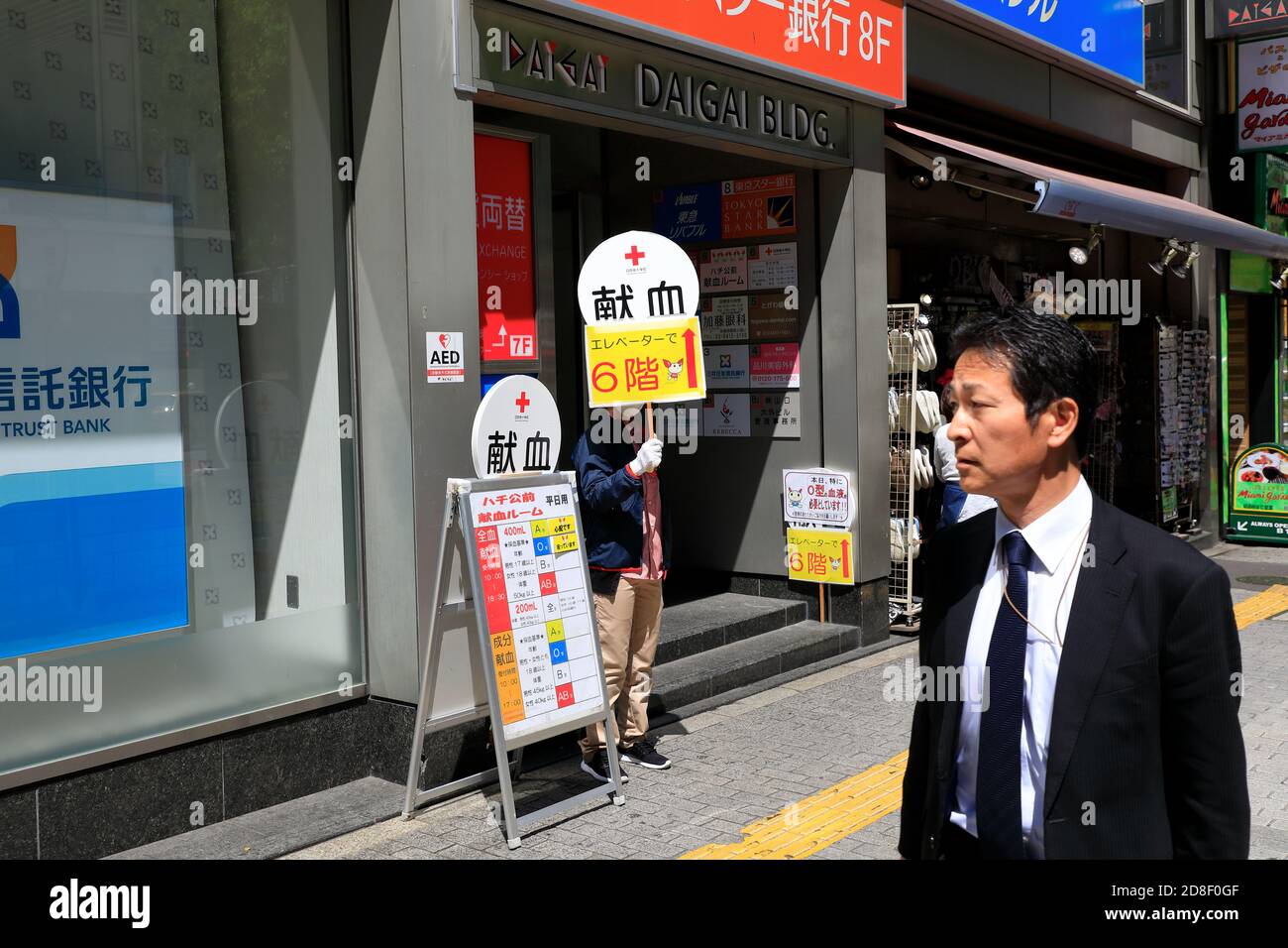 Una persona che tiene un segno che chiede la donazione di sangue vicino l'ingresso di una banca del sangue con un passaggio pedonale By.Shibuya.Tokyo.Japan Foto Stock