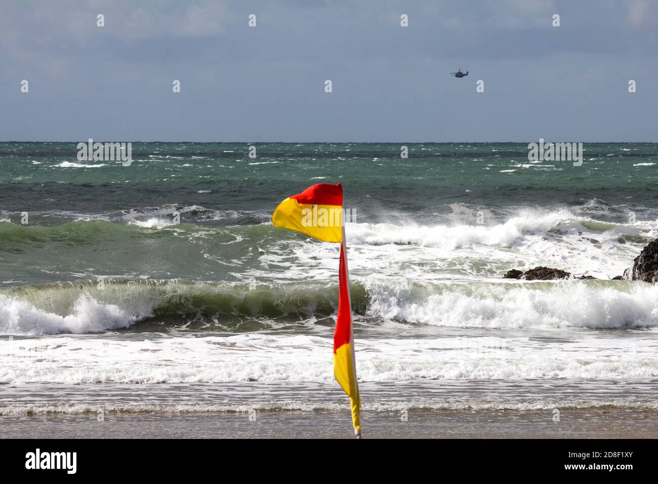 Infrangere le onde sulla spiaggia di Poldhu, Poldhu Cove, Cornovaglia. Una baia popolare per i surfisti. Bandiera di allarme RNLI nel vento con un elicottero in lontananza. Foto Stock