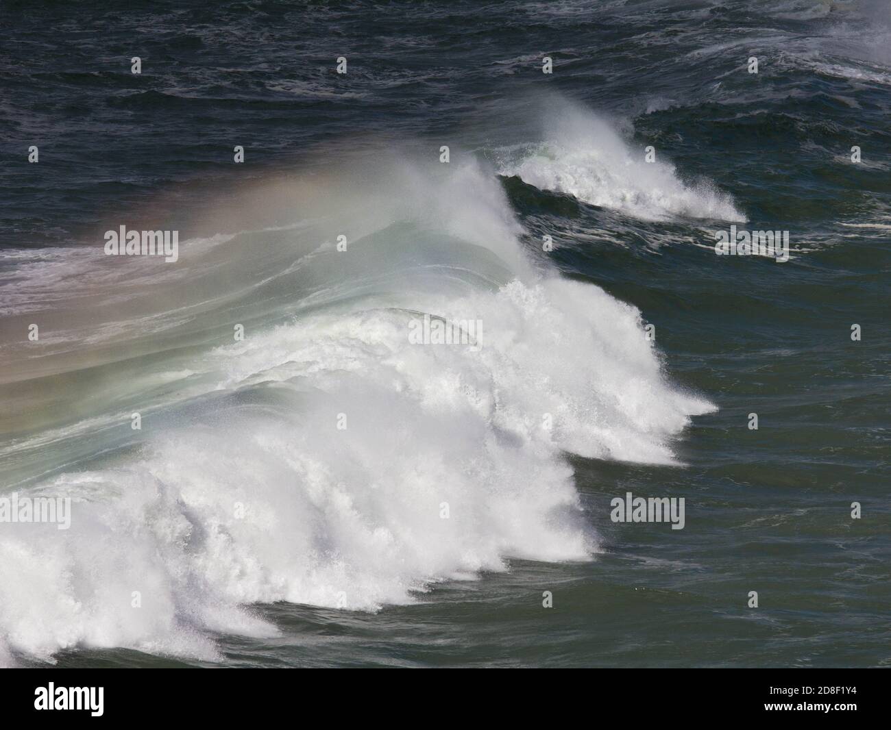 Infrangere le onde con un arcobaleno nello spray spazzato dal vento. Vicino alla spiaggia di Poldhu, Poldhu Cove, Cornovaglia. Un'insenatura popolare per surfisti e principianti di surf. Foto Stock