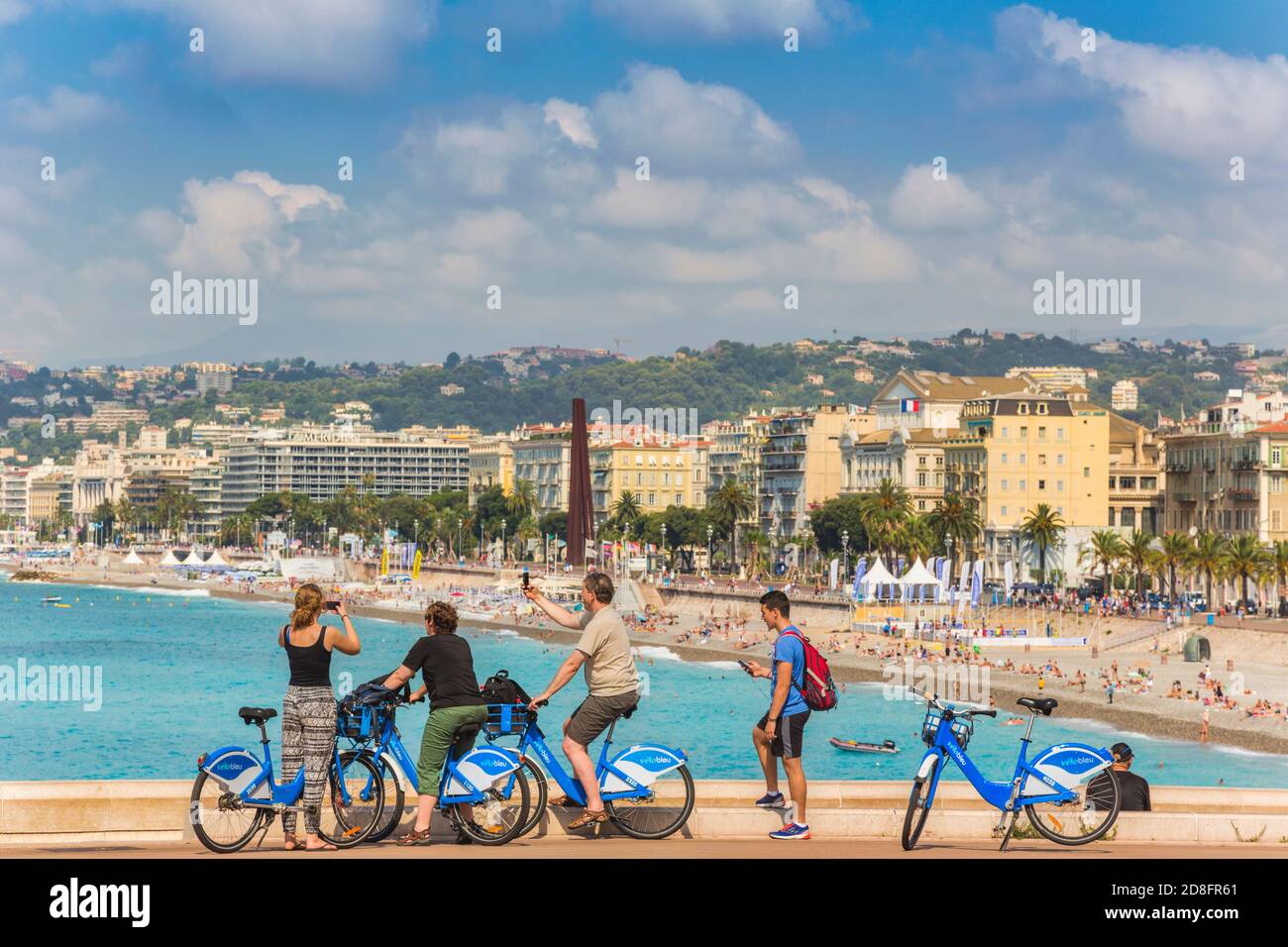 Nizza, Costa Azzurra, Costa Azzurra, Francia. I ciclisti scattano fotografie sui loro smartphone della spiaggia di fronte al lungomare di Nizza. Foto Stock