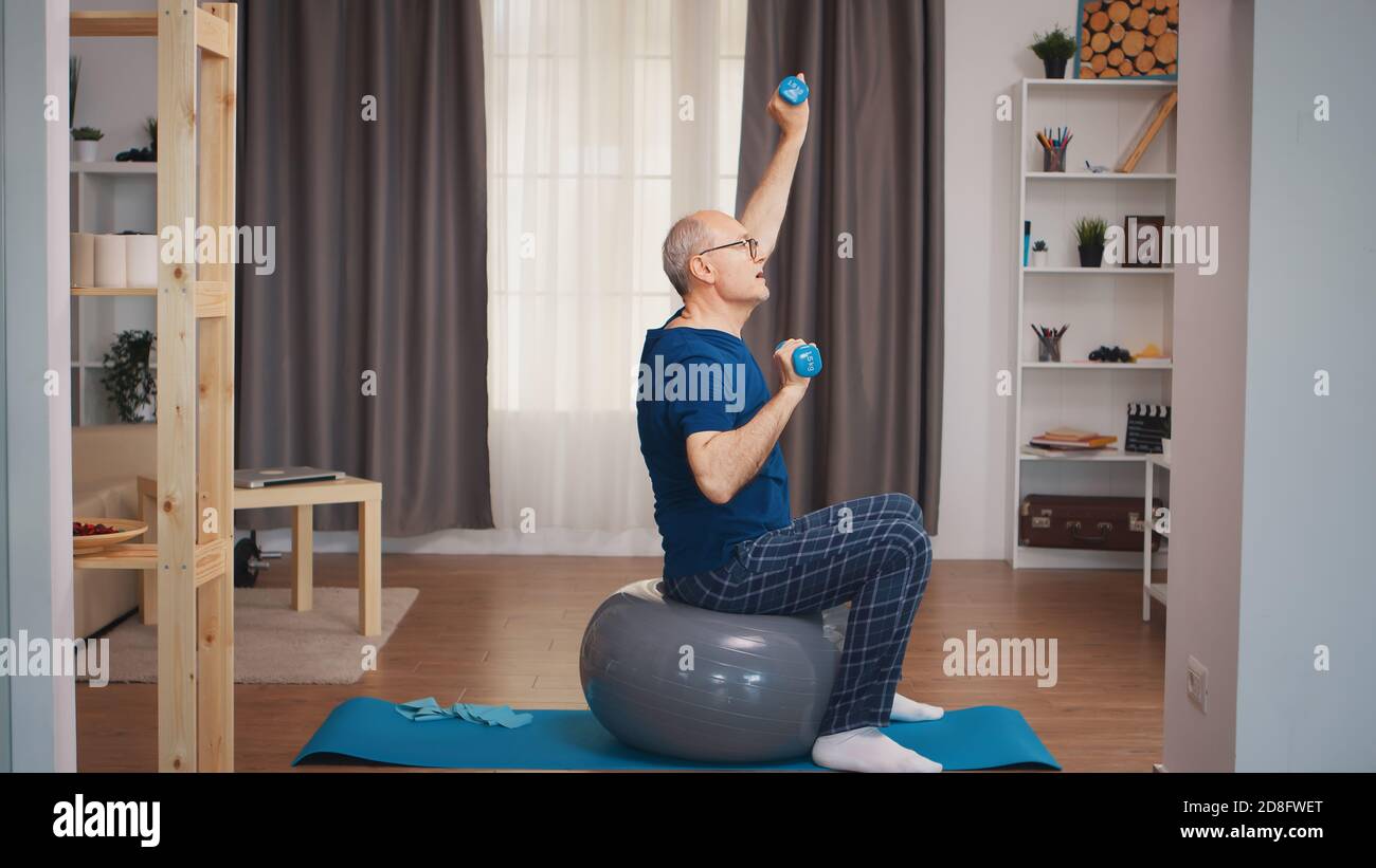 Uomo anziano con vitalità esercitarsi usando i manubri nel soggiorno. Anziano pensionato sano addestrando sport di salute a casa, esercitandosi attività di idoneità all'età anziana Foto Stock
