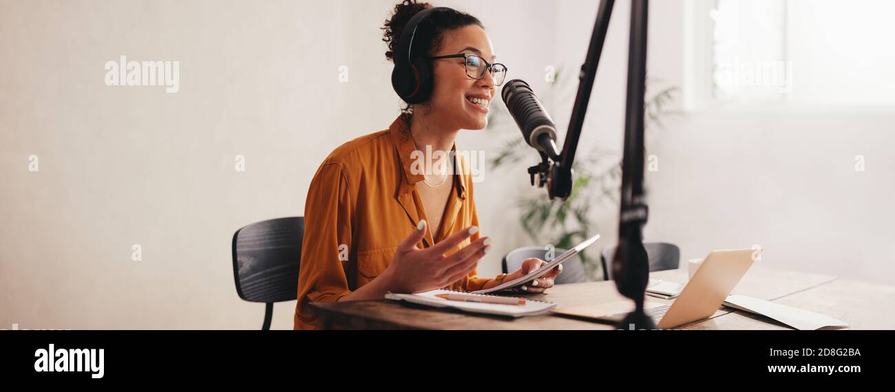 Donna che registra un podcast sul suo computer portatile con cuffie e un microscopio. Podcaster femminile che crea podcast audio dal suo studio di casa. Foto Stock