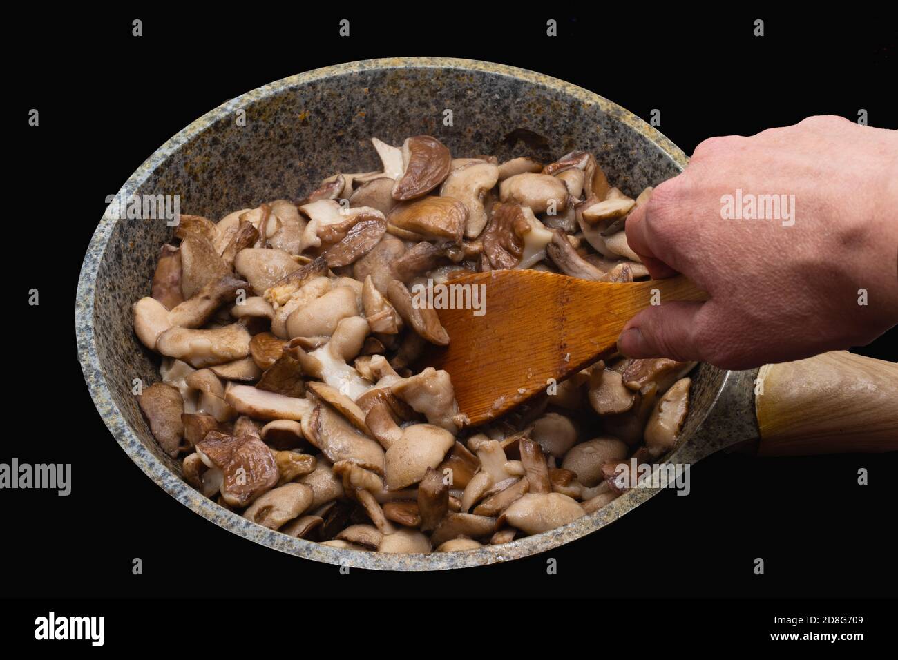 Una mano di donna si gira su un cucchiaio di legno di fritto o i funghi stufati in una padella su un nero sfondo Foto Stock