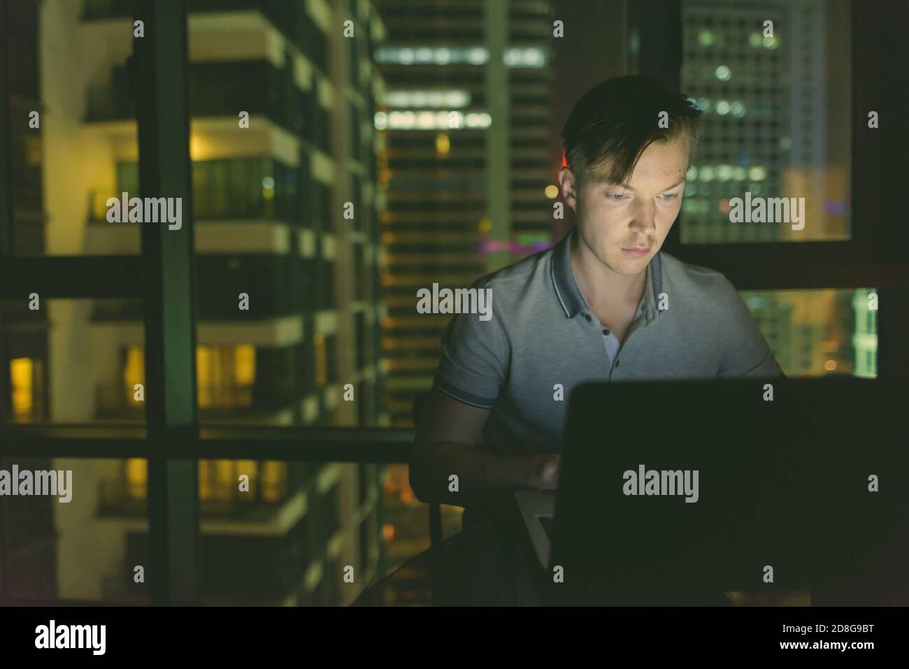 Giovane bell'uomo che usa il laptop contro la finestra di vetro con vista della città di notte Foto Stock