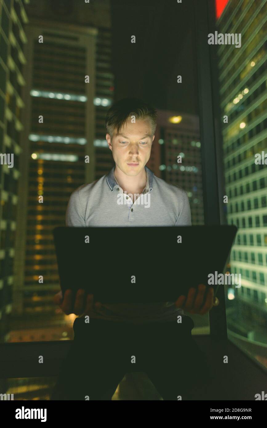 Giovane bell'uomo che usa il laptop contro la finestra di vetro con vista della città di notte Foto Stock