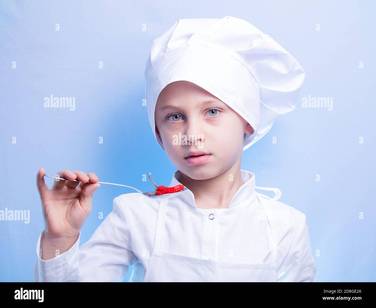 un ragazzo di 6 anni in tuta e il cappellino dello chef sorride alla macchina fotografica e tiene un peperoncino caldo in un cucchiaio durante una degustazione. il concetto di cucina di un bambino Foto Stock