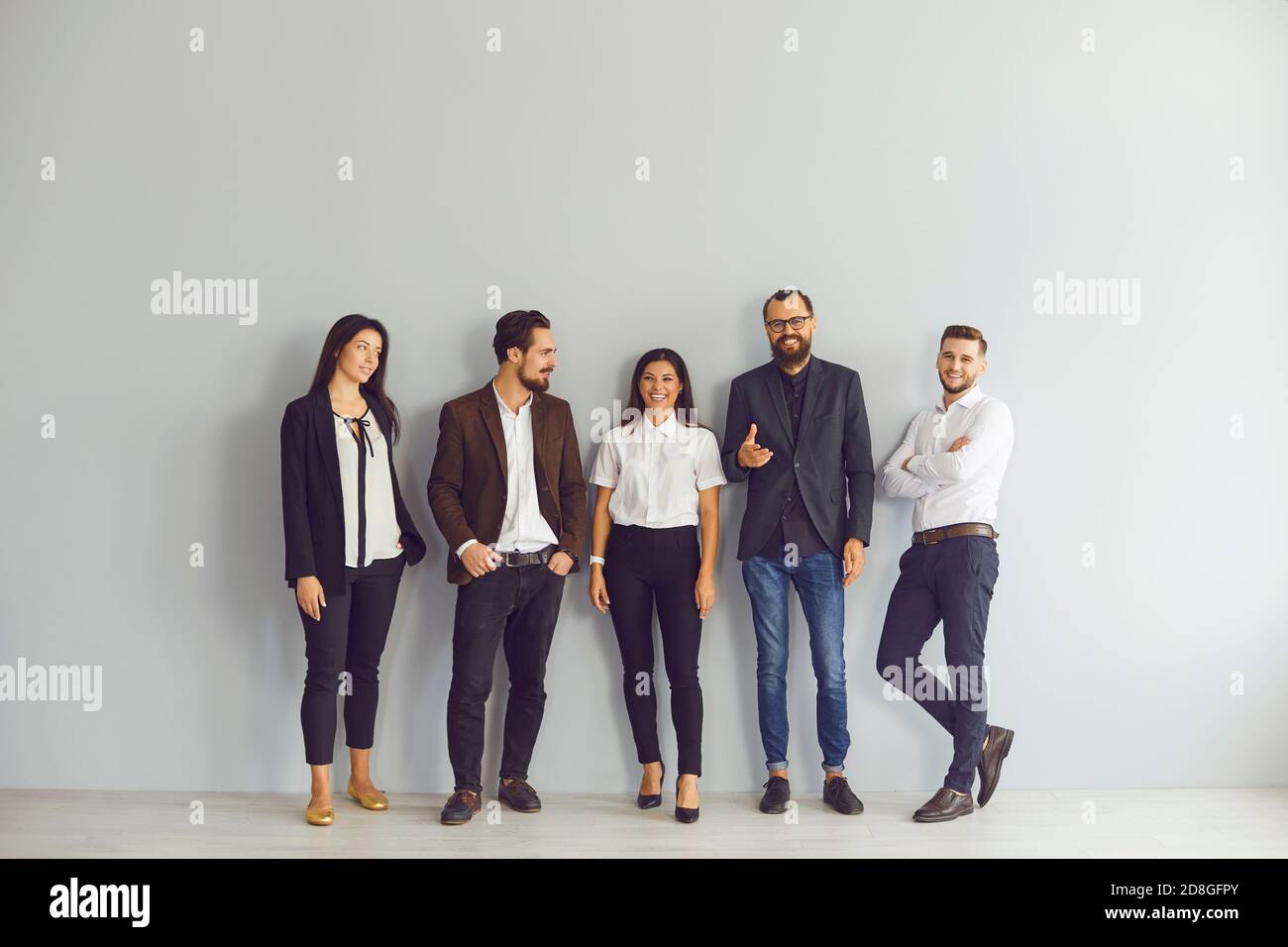 Gruppo di giovani uomini d'affari felici che si trovano vicino alla parete dello studio guardando la fotocamera Foto Stock