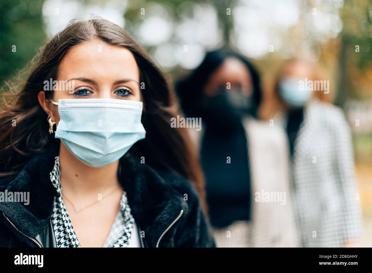 mettere a fuoco in primo piano su una giovane donna in piedi all'aperto in coda e indossare una maschera protettiva con l'altro defocused di etnici misti Foto Stock