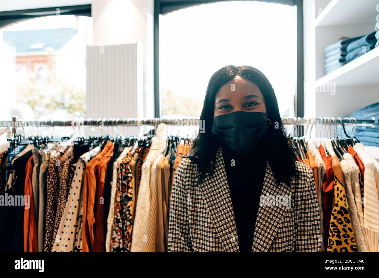 ritratto di una bella donna nera che indossa una maschera facciale nel suo negozio di moda. Un'allegra lavoratrice che si trova di fronte alla fotocamera in boutique Foto Stock