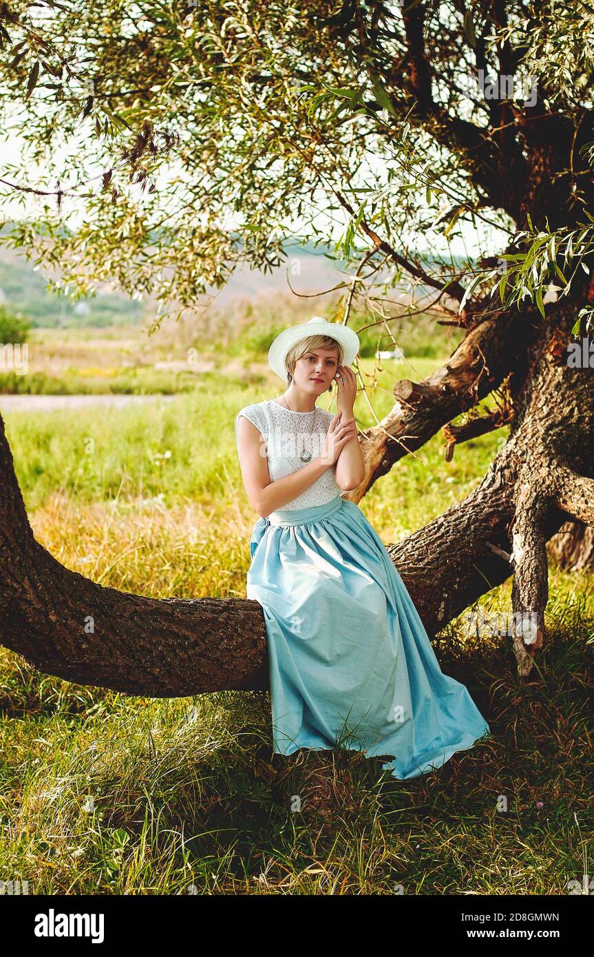 Giovane elegante donna felice con cappello in abiti vintage camminando da sola nella natura in campo verde. Foto Stock