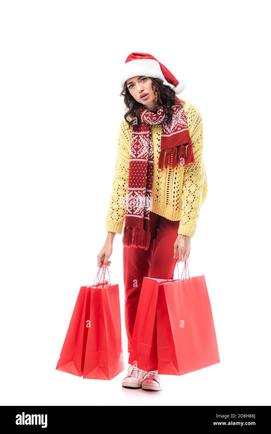 stanca giovane donna in cappello santa e sciarpa con ornamento tenere le borse per la spesa rosse isolate su bianco Foto Stock