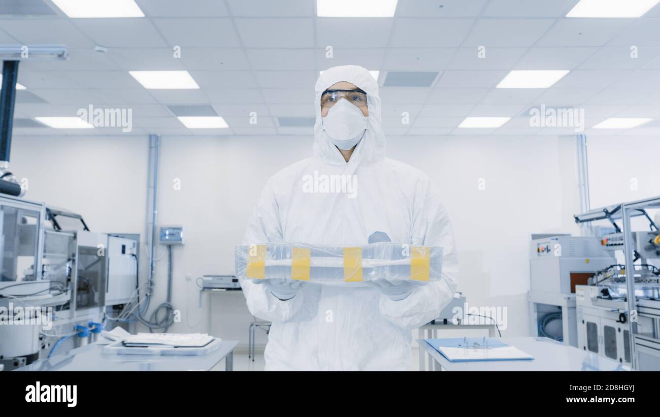 Lo scienziato in tuta protettiva trasporta la custodia con il prodotto finito attraverso il laboratorio. Struttura con macchinari industriali moderni. Produzione di prodotti Foto Stock