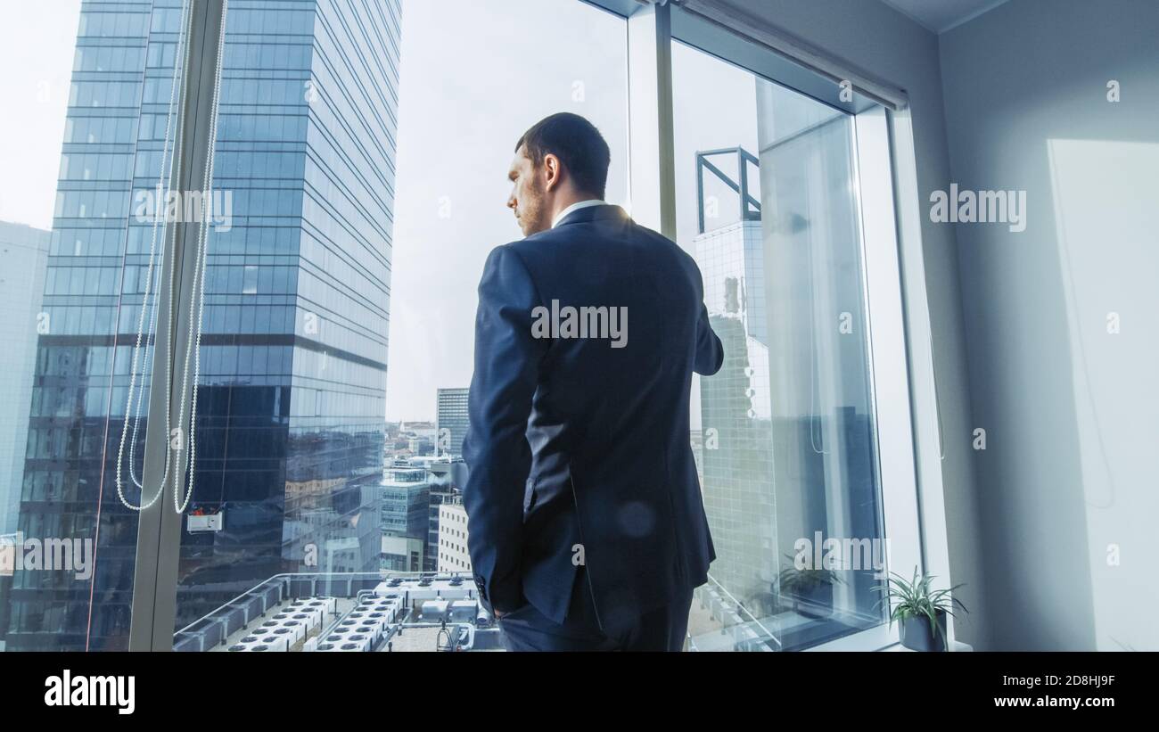 Uomo d'affari pensieroso che indossa vestito in piedi nel suo ufficio, guardando fuori dalla finestra e contemplando il prossimo contratto di grande impresa. Città maggiore Foto Stock