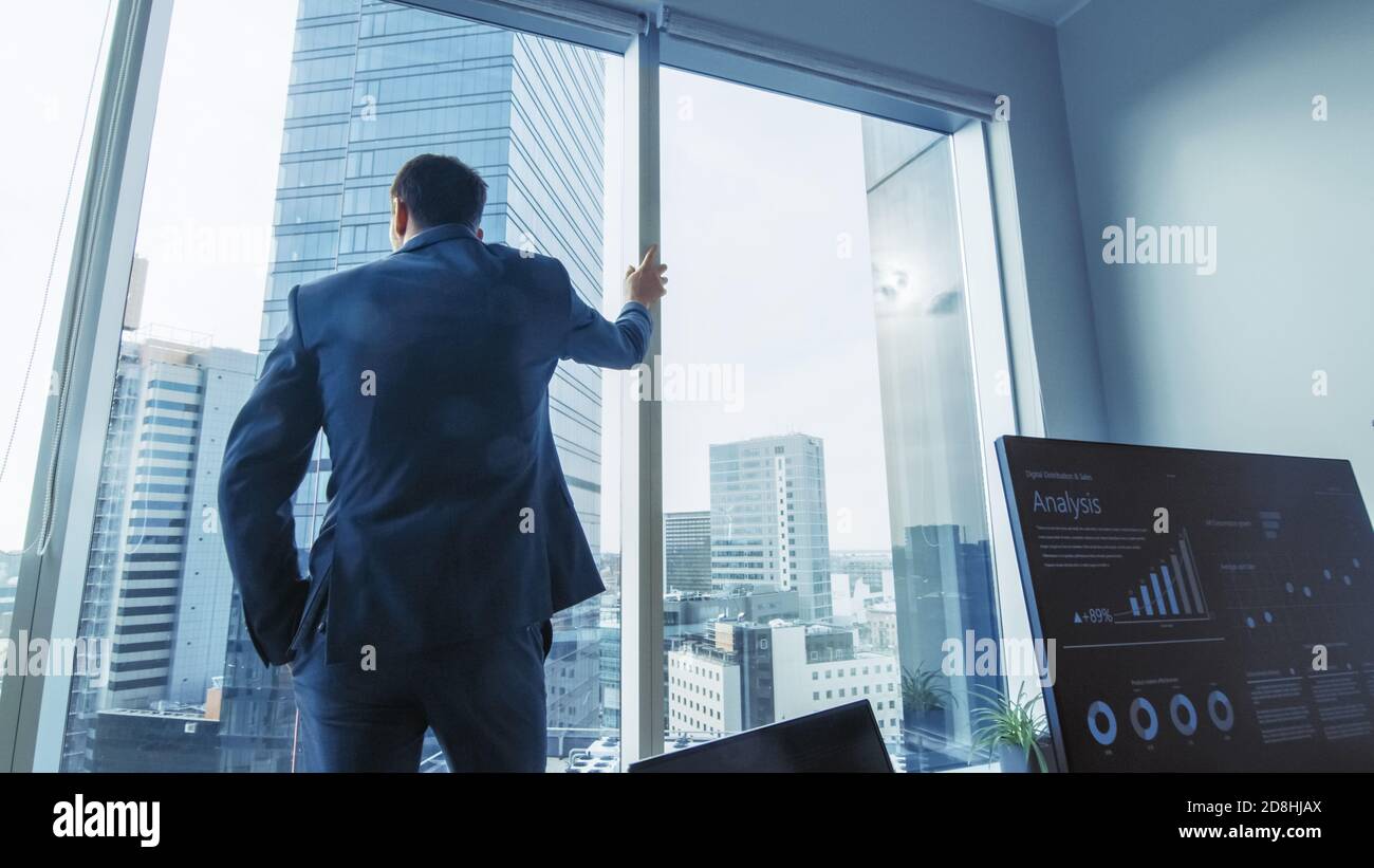 Uomo d'affari pensieroso che indossa vestito in piedi nel suo ufficio, guardando fuori dalla finestra e contemplando il prossimo contratto di grande impresa. Città maggiore Foto Stock