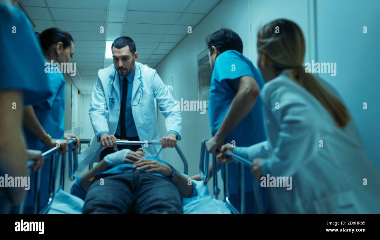 Medici, infermieri e chirurghi del reparto di emergenza spostano gravemente il paziente ferito che giace su un Gurney attraverso i corridoi dell'ospedale. Personale medico in un Foto Stock