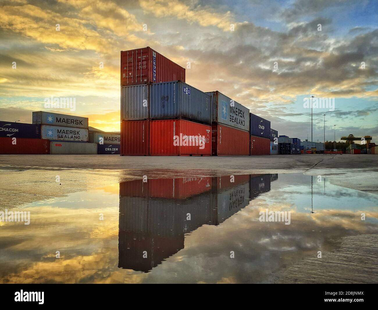 Contenitori di spedizione e scatole di carico impilati su un banchina deserta al tramonto riflesso in acqua e spazio copia Foto Stock