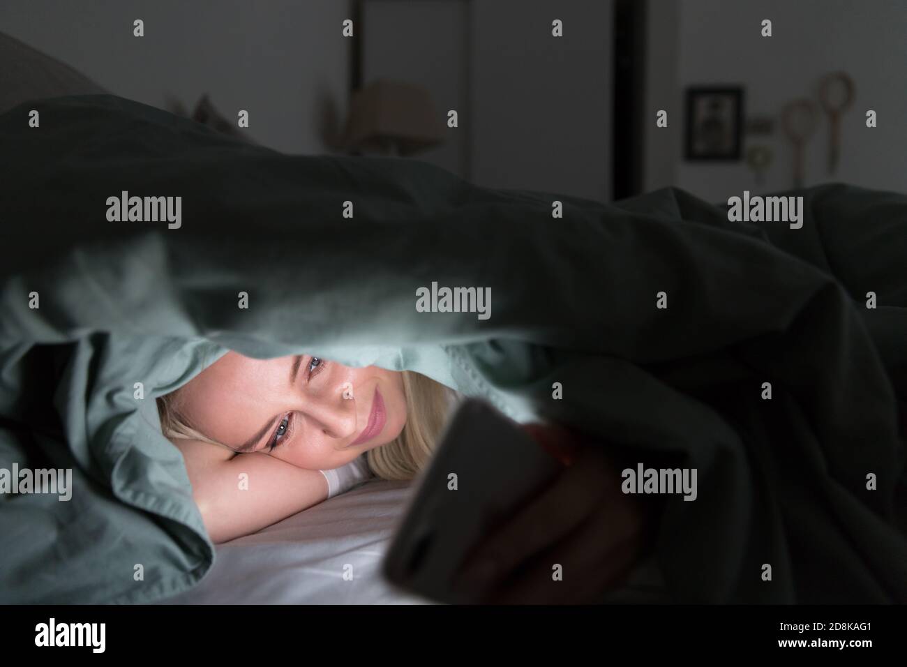 Giovane donna allegra sdraiata a letto sotto la coperta utilizzando il telefono cellulare a tarda notte, non può dormire. Insonnia, nomofobia, concetto di dipendenza da Internet Foto Stock