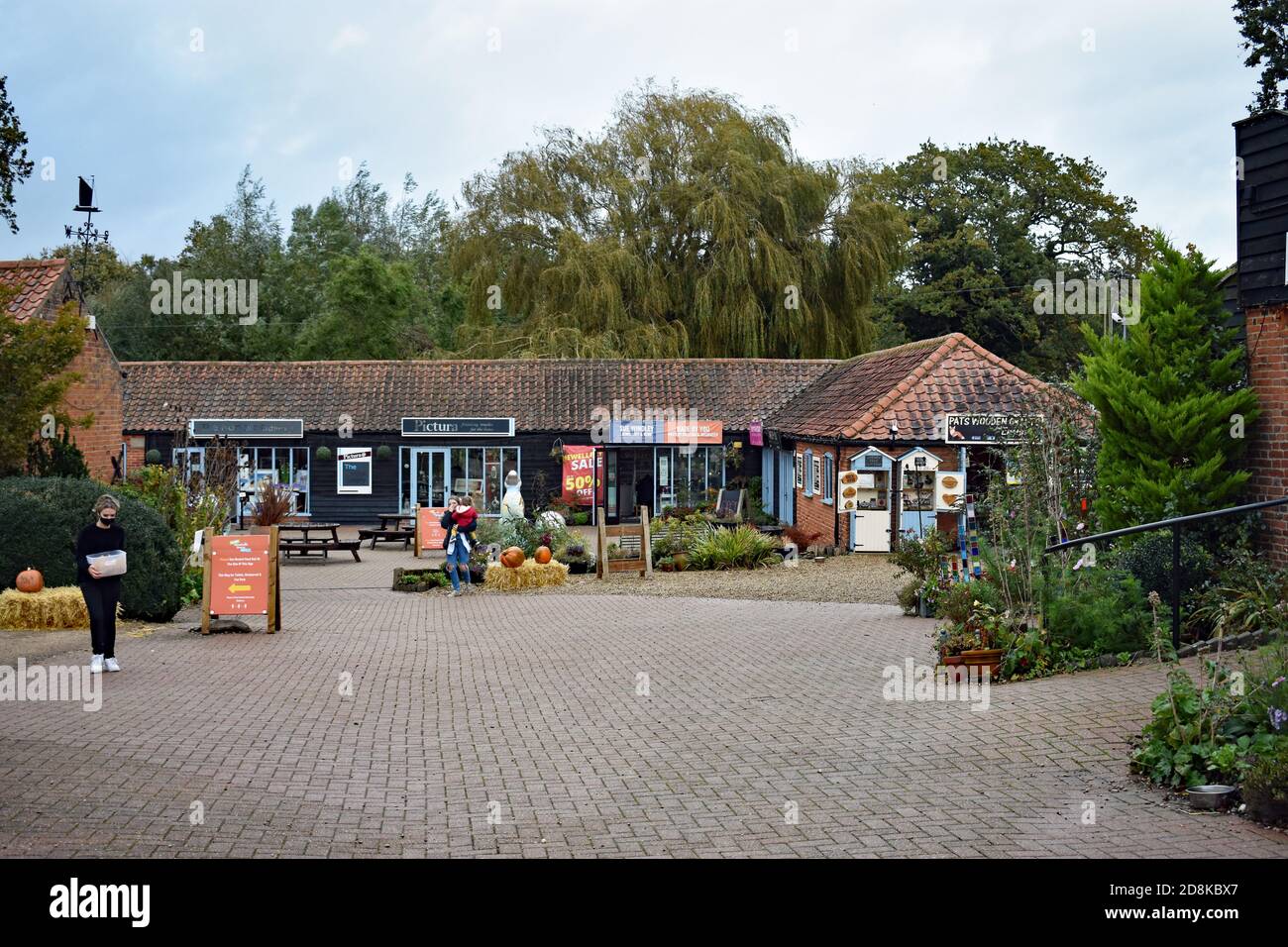 Una donna che indossa una maschera porta una scatola di plastica attraverso il cortile pavimentato in mattoni a Wroxham Barns, Norfolk, UK. I fienili contengono boutique. Foto Stock