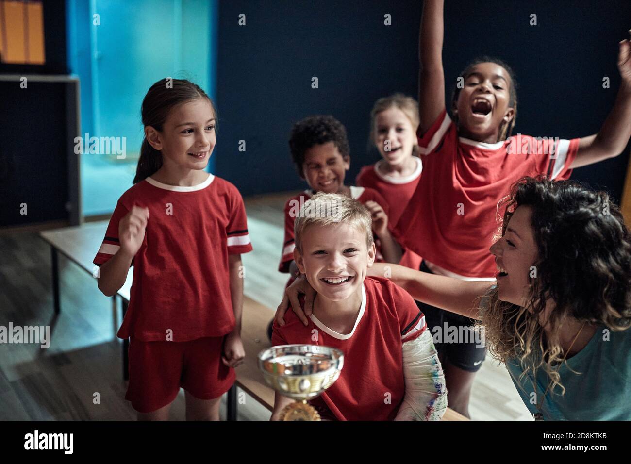 La squadra dei bambini e la allenatrice femminile presso l'armadietto in posa con il trofeo vinto. Sport di squadra per bambini Foto Stock