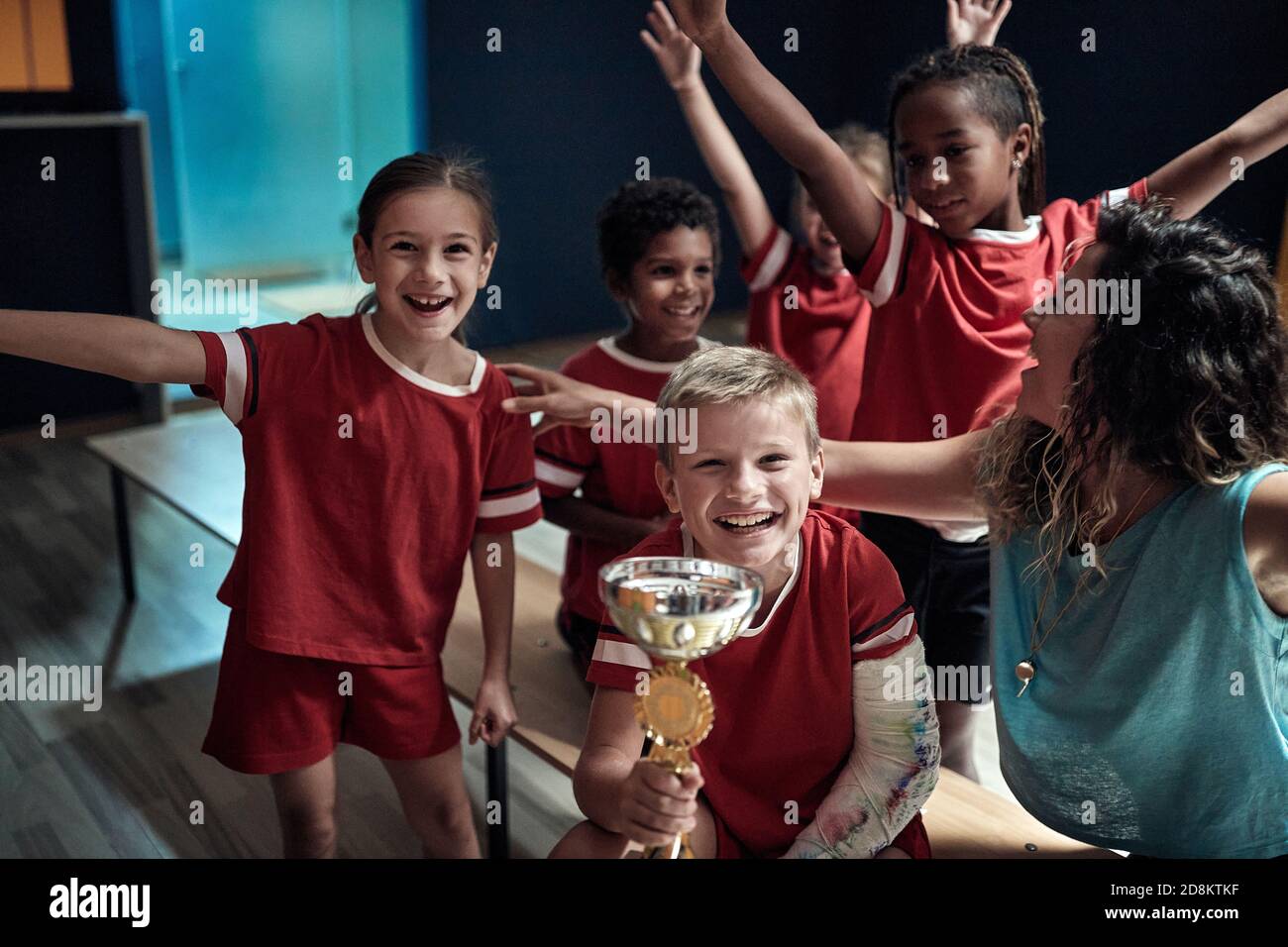 La squadra dei bambini e la allenatrice femminile si sono eccitate presso l'armadietto per il trofeo vinto. Sport di squadra per bambini Foto Stock