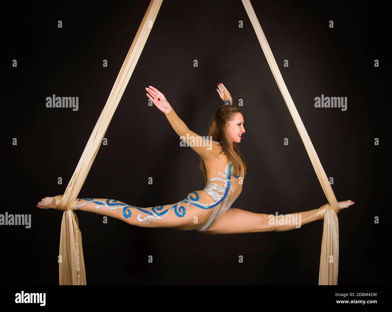 Una donna snella in una tuta beige esegue ginnastica ed esercizi circensi sulla seta dorata. Riprese in studio su sfondo scuro, immagini isolate. Foto Stock