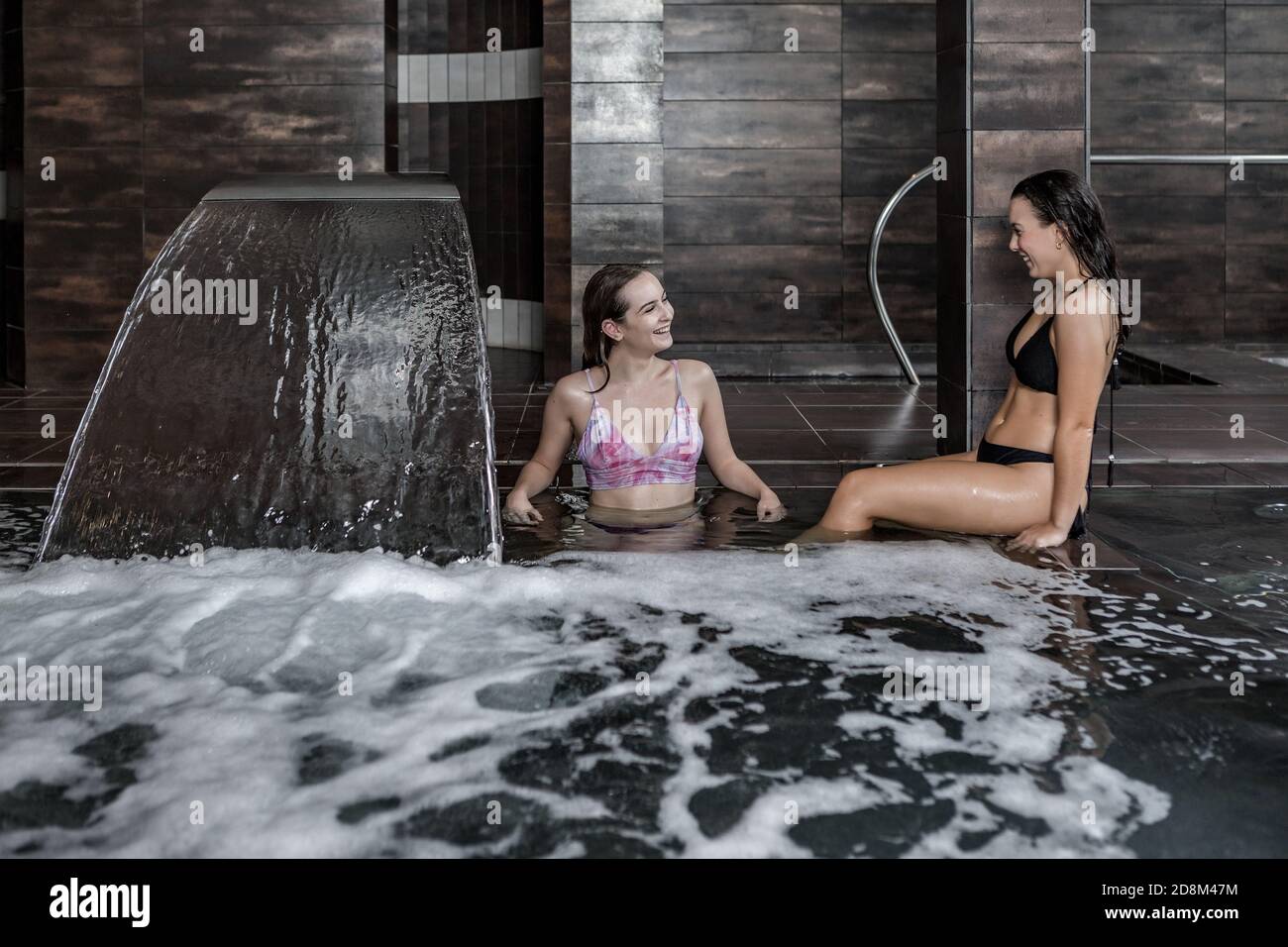 Allegre amiche che parlano e ridono mentre si gustano i massaggi idro in piscina con acqua schiumosa e cascata nella moderna spa centro Foto Stock
