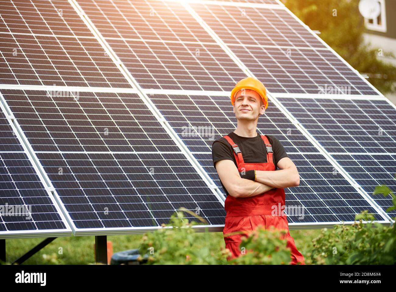 Orgoglioso giovane lavoratore sorridente soddisfatto del suo lavoro di successo con le sue braccia incrociate, sullo sfondo stazione solare. Concetto: Energia rinnovabile, tecnologia, elettricità, servizio, energia verde. Foto Stock