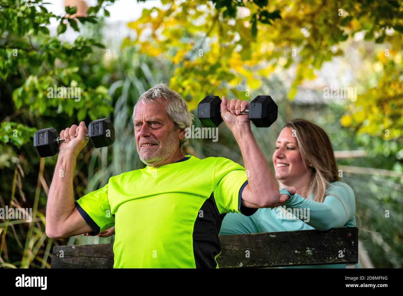 Un uomo alla fine degli anni sessanta solleva i pesi all'aperto con l'aiuto di una giovane personal trainer femminile. Foto Stock