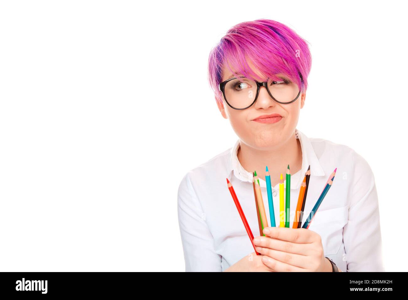 Giovane donna grumpia con acconciatura rosa che indossa occhiali e camicia tenere insieme di matite colorate e guardare lontano in delusione isolato su wi Foto Stock