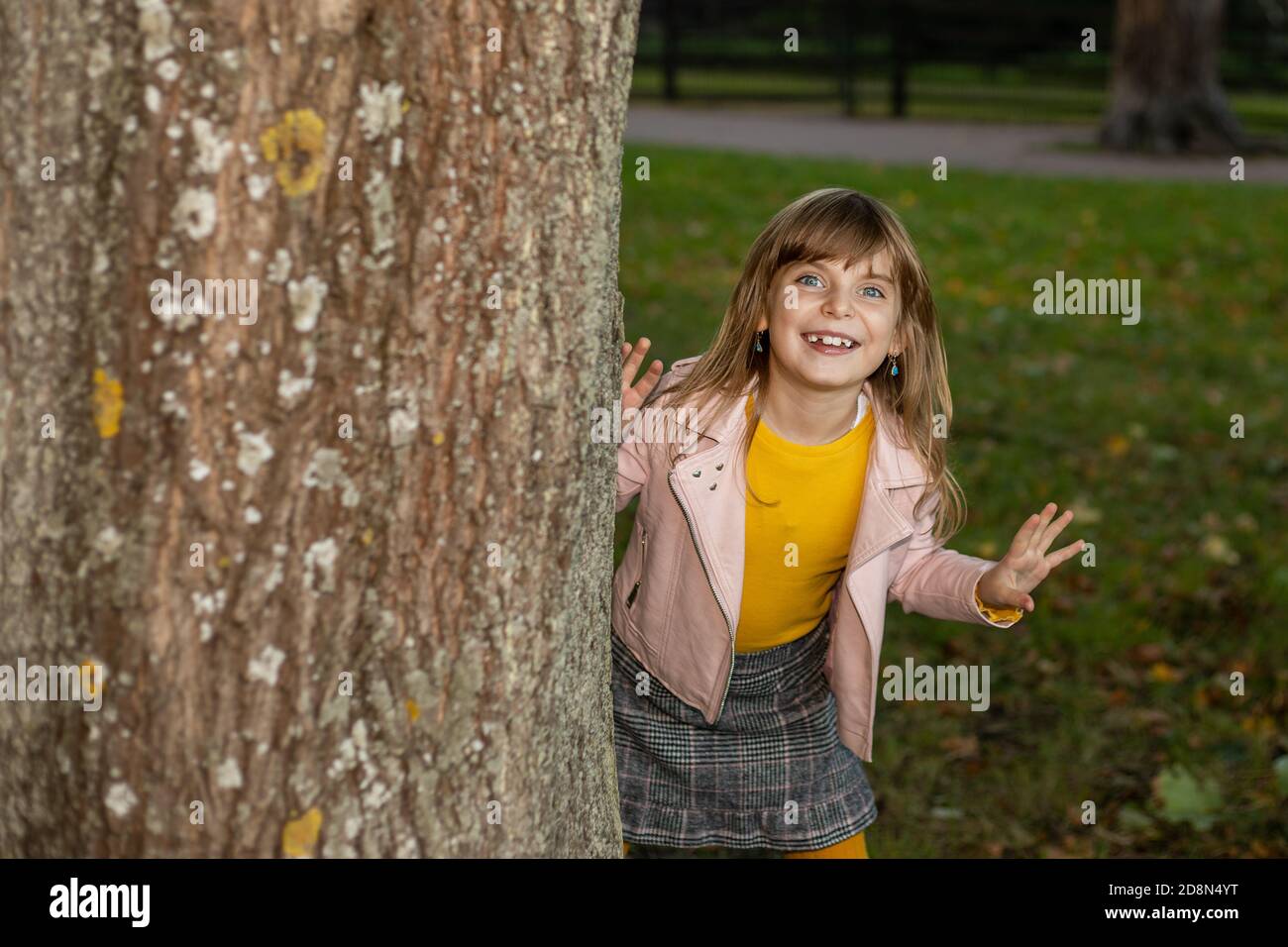 Toothless divertente ragazza carina di capretto che gioca nascondino e cerchi nel parco autunnale. Si affaccia da dietro un albero con un sorriso divertente e ride. Concetto stagionale Foto Stock
