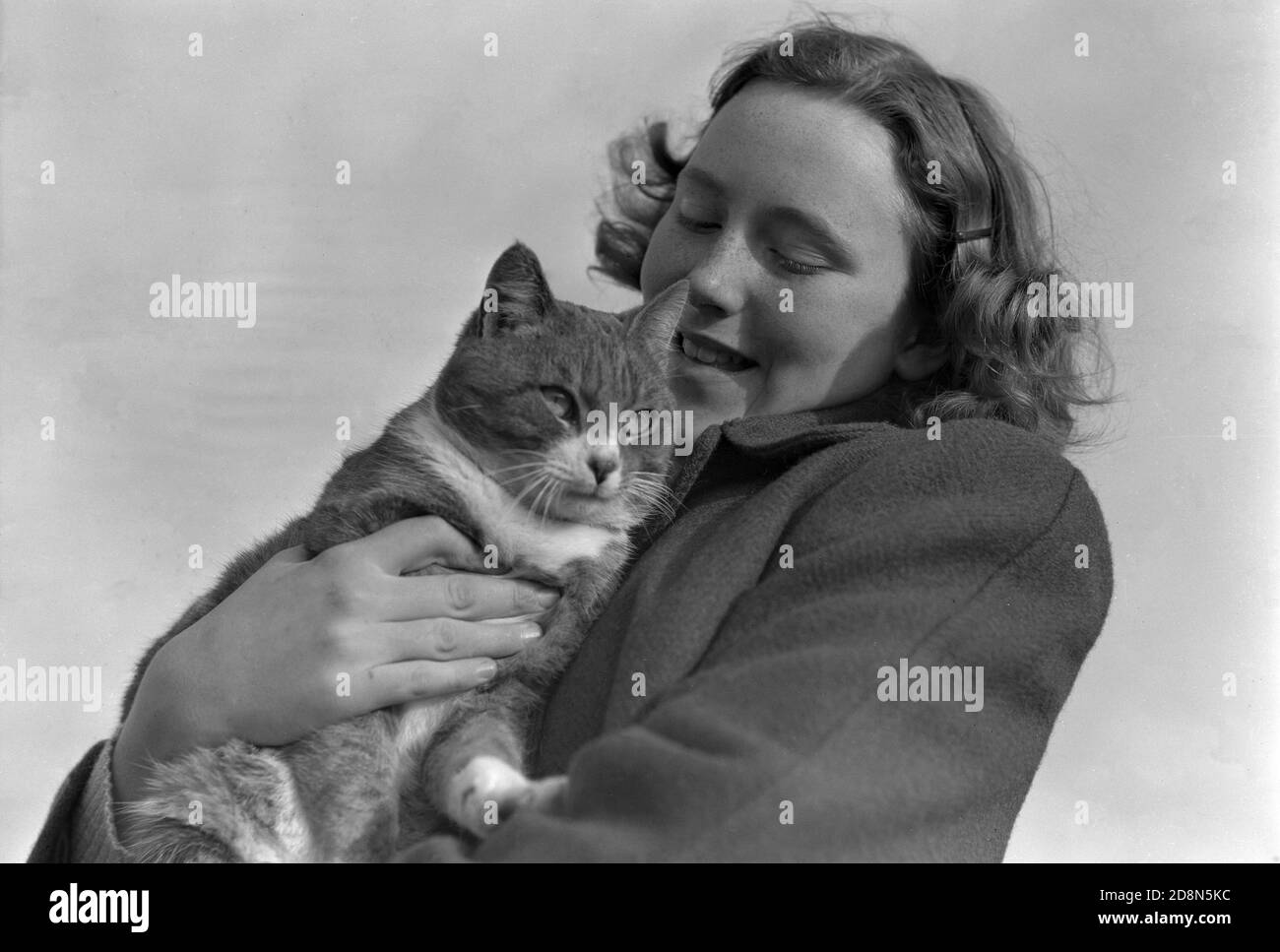1950s, storica, una giovane donna attraente in piedi all'aperto cocdling il suo gatto dell'animale domestico. Ci è molta prova che gli animali domestici di stroking o cocdling quali i gatti ed i cani ha benefici significativi di salute e di umore-miglioramento per i loro proprietari. Foto Stock