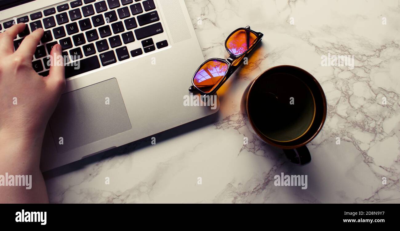 Scrivania con un computer portatile, come vetri di blocco blu, e tazza con la mano sulla tastiera. Foto Stock