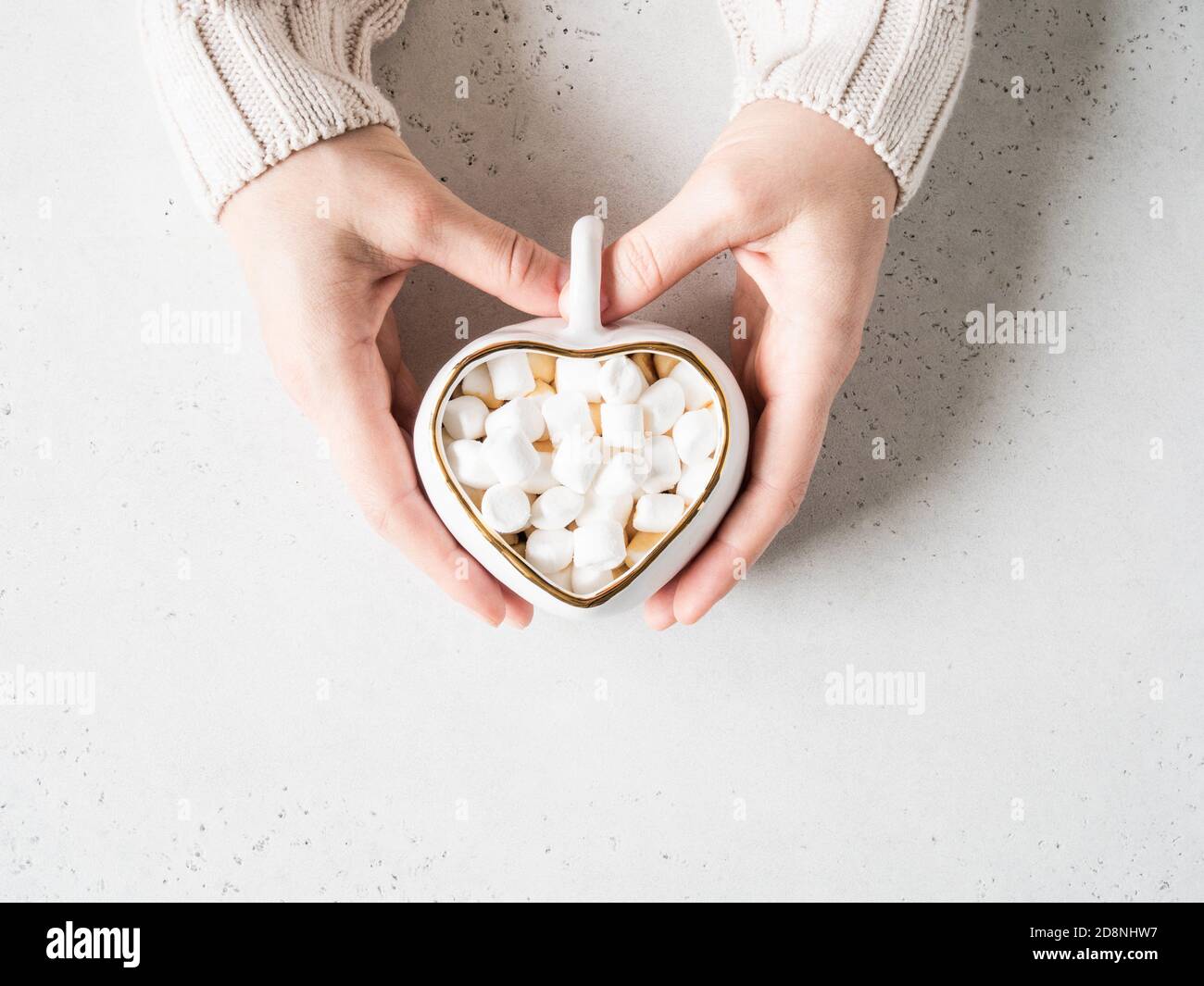 Le mani femminili tengono la tazza bianca di ceramica sotto forma di cuore con la bevanda calda e il marshmallows su sfondo chiaro. Vista dall'alto. Spazio di copia Foto Stock