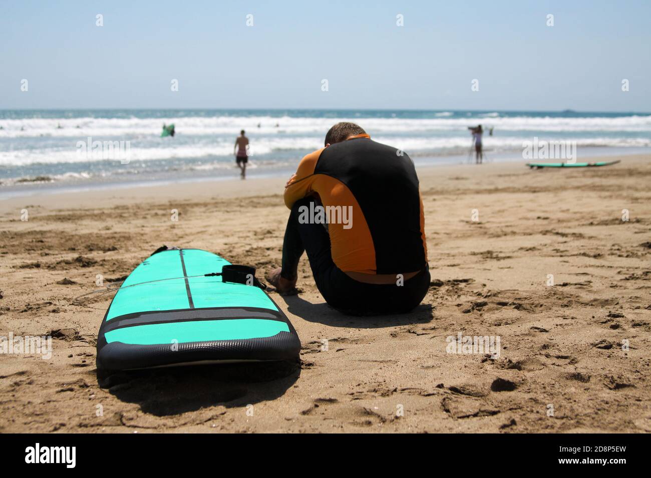 Surfer principiante in un costume da bagno è seduto sulla sabbia bagnata sull'oceano. La sua testa è piegata su mani piegate. Accanto c'è una tavola da surf. Surfista Foto Stock