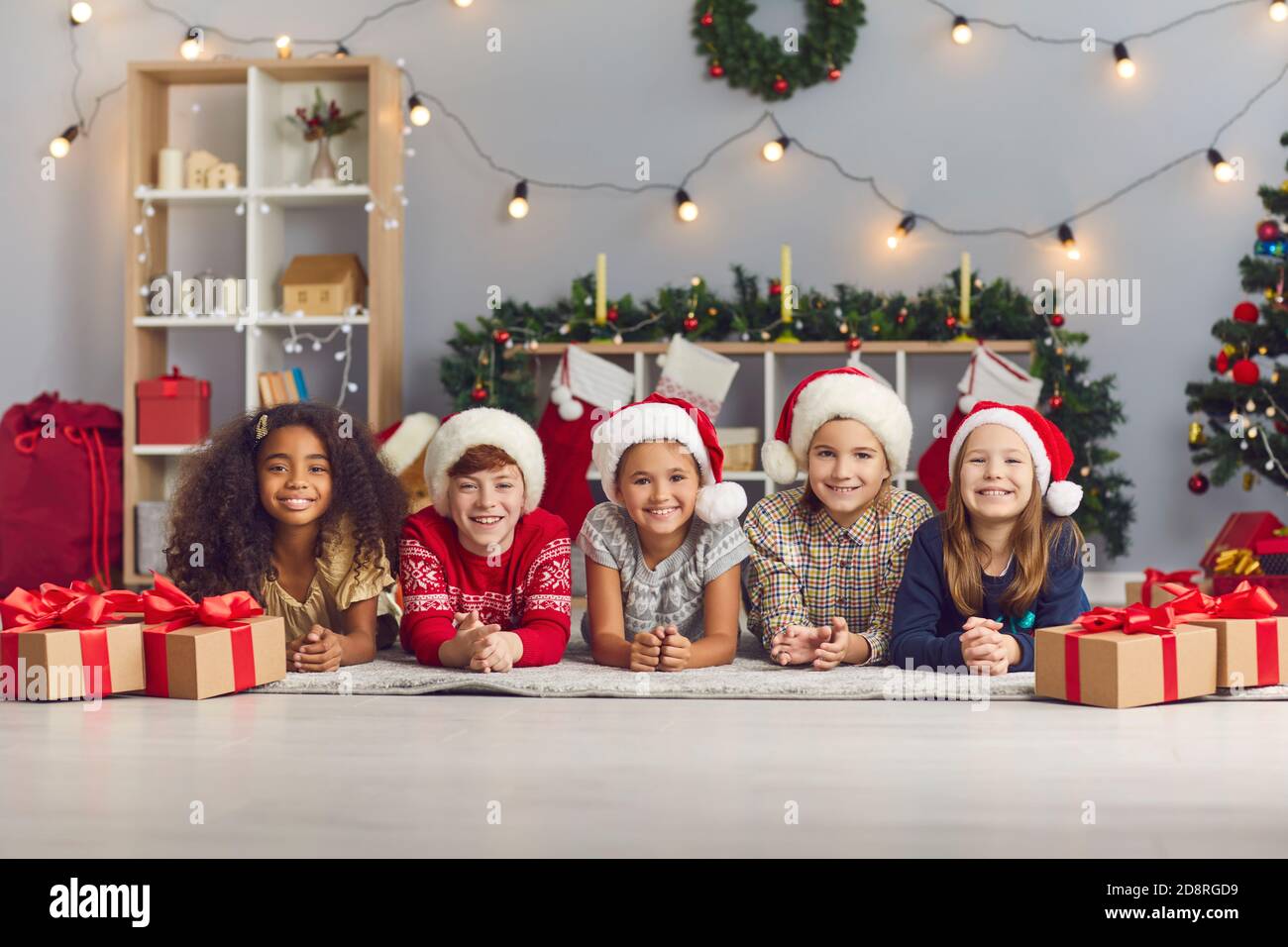 Gruppo di bambini felici a Santa cappelli che guardano la macchina fotografica sdraiato su un piano in un accogliente soggiorno con regali Foto Stock